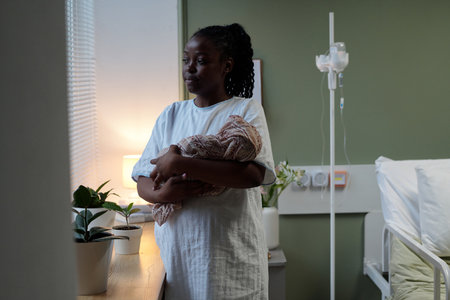 Mother standing by hospital room window holding newborn baby wrapped in blanket gazing thoughtfully. Room features hospital bed, IV stand, potted plant, and soft natural light coming through windowの写真素材