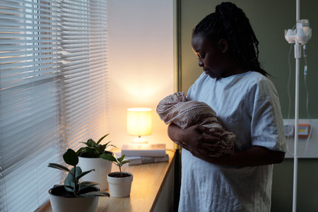 African American mother holding newborn baby while standing by window in hospital room with potted plants and soft lamp light creating serene ambianceの写真素材