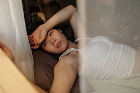 Young Chinese woman suffering from headache and hangover after home party resting on bed by window on sunny morningの写真素材
