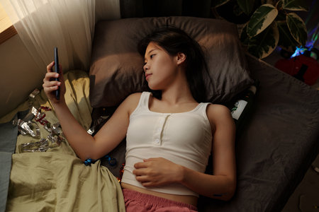 High angle of young Chinese woman with mobile phone resting on bed after booze party and taking selfie or textingの写真素材