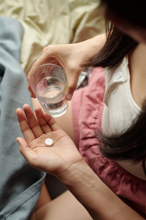 High angle of hands of teenage girl in homewear sitting on bed in the morning and taking pill healing symptoms of hangoverの写真素材