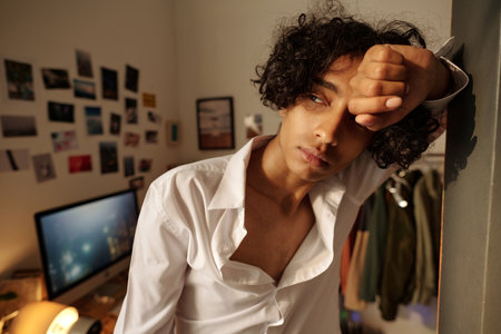 Sick guy in white shirt looking aside through window while standing against wall and suffering from hangoverの写真素材