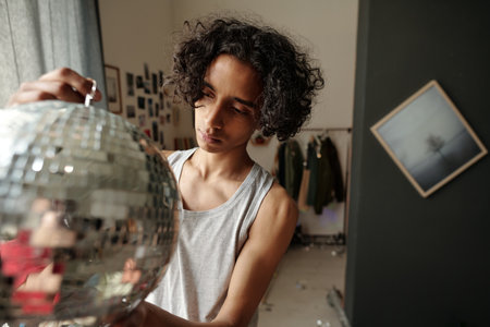 Teenage guy with hangover symptoms looking at sparkling disco ball while cleaning domestic room...