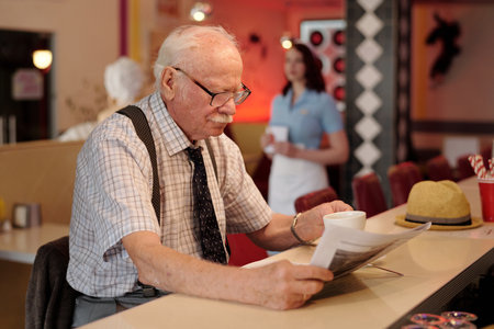 Senior man sitting at counter reading newspapers with waitress in background serving a customer Standing nearby are stools and mugs creating a comfy environmentの写真素材