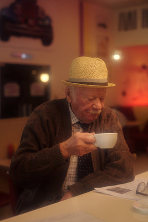 Elderly man sipping coffee at a cozy cafe with warm lighting and a relaxing atmosphere, wearing a brown cardigan and a hat, enjoying a peaceful moment during his dayの写真素材