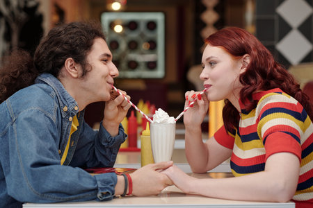 Young couple sharing milkshake in retro-themed diner, both enjoying their time together. Blurred...