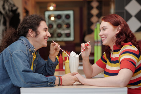 Smiling couple holding hands while enjoying milkshakes at an ice cream shop, both dressed in colorful, casual outfits engaging in cheerful conversation with milkshake and condiments on tableの写真素材