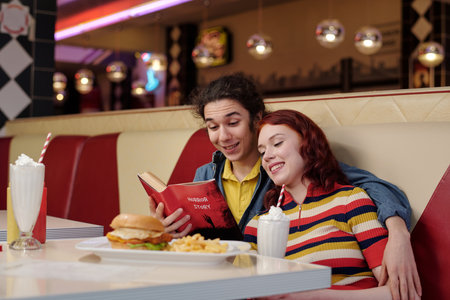 Friends sitting in cozy diner booth while sharing laugh over book and enjoying tasty milkshakes and...