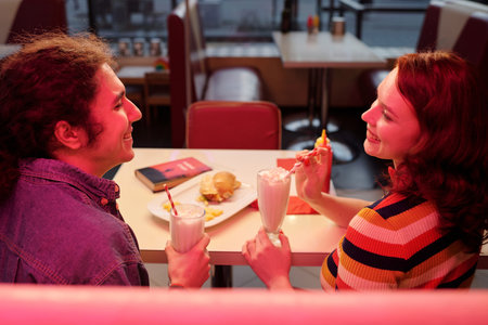 Two people sipping milkshakes and sharing a meal at retro-themed diner, capturing warm ambiance and cheerful interactionの写真素材
