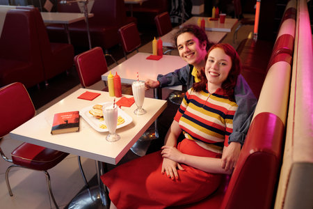 Smiling couple sitting closely in classic retro diner booth enjoying milkshakes and meal, books placed nearby on the table. Decor with red and white upholstery creating vibrant atmosphereの写真素材