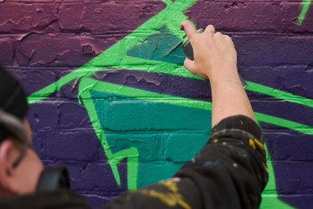 Hand of young unrecognizable man with can of graffiti paint spraying it on brick wall while outlining purple letter or figureの写真素材