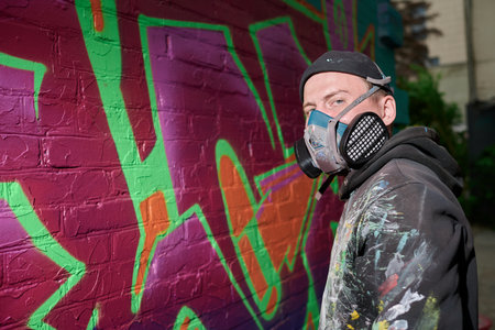 Creative guy in stylish hoodie, beanie and respirator standing by brick wall with graffiti artwork while looking at cameraの写真素材
