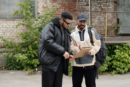 Two men are standing in front of building, discussing documents on a clipboard. One is wearing a black jacket, while other is in beige jacket and cap, both appear focused on their taskの写真素材