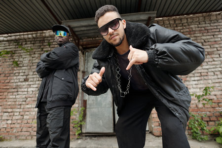 Two men showcasing street fashion with urban backdrop, wearing shades while posing confidently. Displaying a cool and edgy vibe, their attire adds to the modern street styleの写真素材