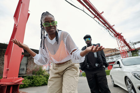 Woman with braids and green sunglasses making a gesture with hands, man in black outfit standing...
