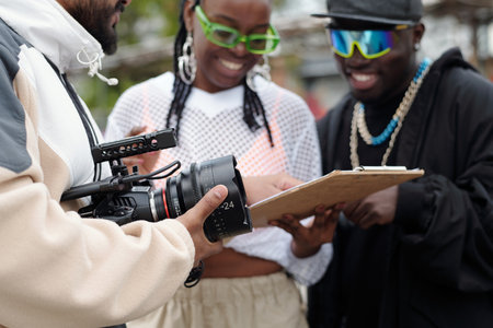 Filming crew holding camera and reviewing script on clipboard during an outdoor film shoot. Team members focused on scene planning without direct eye contact with cameraの写真素材