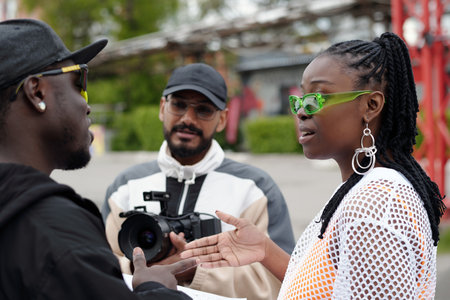 Three people engaging in conversation outdoors with two individuals facing each other and one person holding a camera. One person wears bright green glassesの写真素材