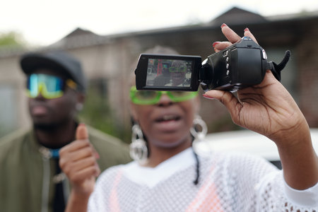 Two people wearing trendy sunglasses and casual outfits, taking selfie with digital camera at outdoor event, smiling and enjoying sunny dayの写真素材