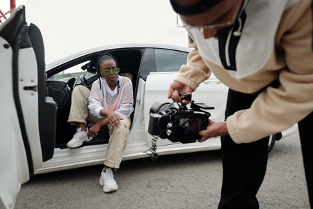 African American woman posing beside white car wearing green sunglasses while being photographed...