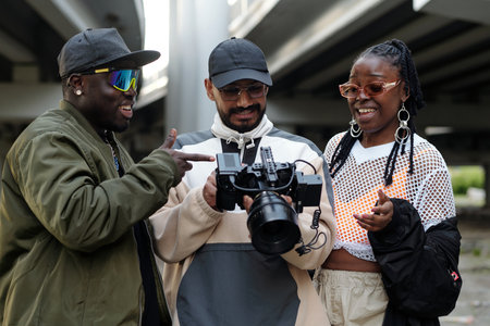 Group of three friends examining camera under bridge with smiles and engaged expressions, reflecting...