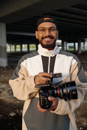 Man wearing glasses and cap smiling while holding professional camera underneath large concrete...
