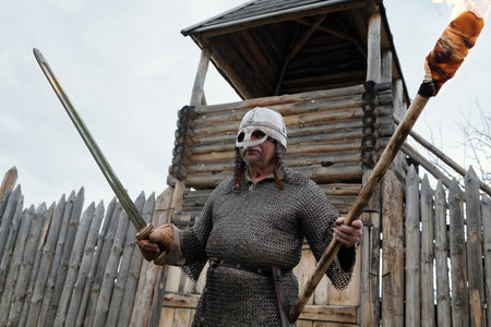 Viking warrior standing in front of wooden fort, holding sword and torch. Wearing helmet and...