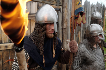 Group of reenactors wearing medieval armor standing in front of wooden fort holding torches while...