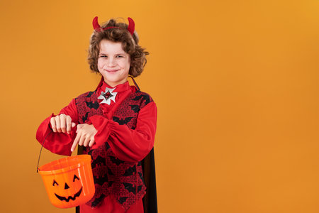 Happy youthful vampire or demon with red horns pointing at bucket for treats and looking at camera...