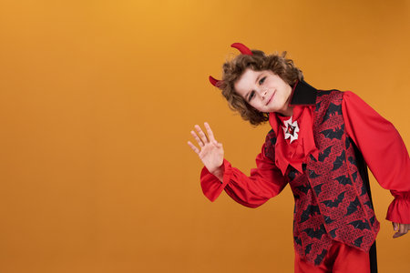 Smiling boy in costume of Halloween demon or vampire waving hand and looking at camera while bending...