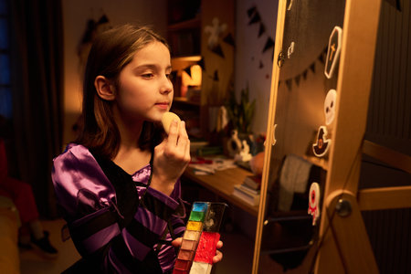 Pretty youthful girl in witch dress putting grease paint on her face with sponge while standing in front of mirror in the eveningの写真素材