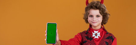 Panorama of boy in Halloween vampire costume looking at camera while holding smartphone with blank...