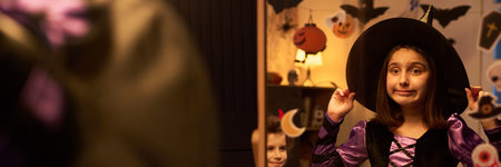 Panorama of mirror reflection of cute little witch in fancy dress and black hat getting ready for Halloween celebration at homeの写真素材