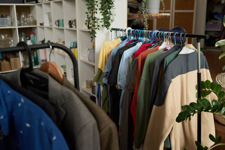 Two racks with secondhand attire standing in front of camera in thrift shop selling clothes, kitchenware, accessories and other stuffの写真素材