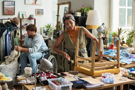 Young female worker of modern secondhand shop hitting part of used wooden chair with hammer while...