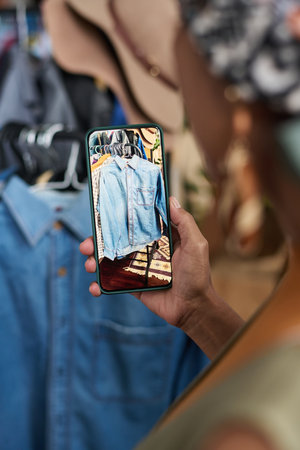Focus on hand of young woman holding smartphone with secondhand denim shirt on screen while taking...