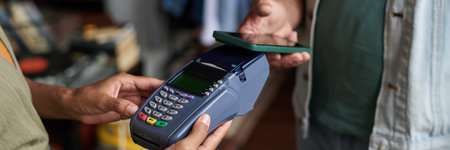 Header of hands of unrecognizable female clerk of thrift shop holding payment terminal in front of male buyer with smartphoneの写真素材