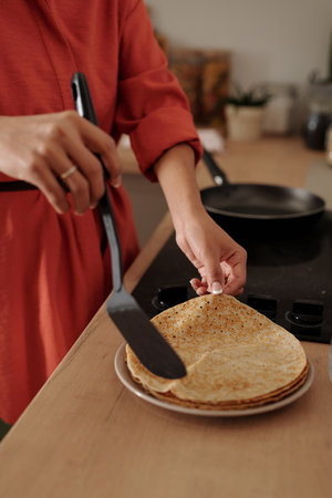 Person wearing red sleeve holding spatula, making crepes on kitchen counter with frying pan, with...