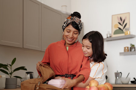 Mother and daughter enjoying time in kitchen, packing lunch in backpack with joy and affection, surrounded by cozy home decor and kitchen essentialsの写真素材