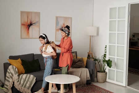 Mother braiding daughters hair with headphones on, creating a bonding moment in cozy living room with modern decor and warm lightingの写真素材