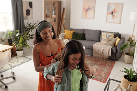 Smiling mother assisting daughter with backpack, standing in modern living room filled with plants...