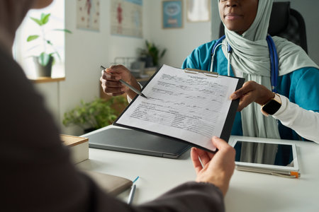Two Muslim women in office environment, one taking notes on clipboard while consulting another. Papers, devices and plants create professional and calm settingの写真素材