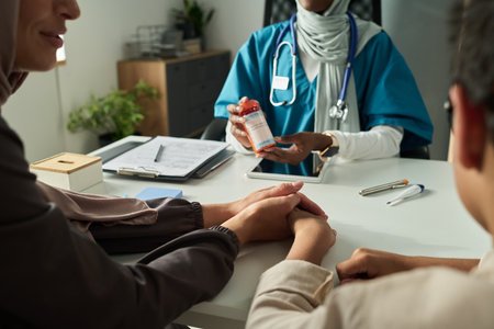 Muslim doctor in blue scrubs explaining medication to patient, holding pill bottle. Patient sitting across table, engaged in conversation with medical professional, sharing concernsの写真素材