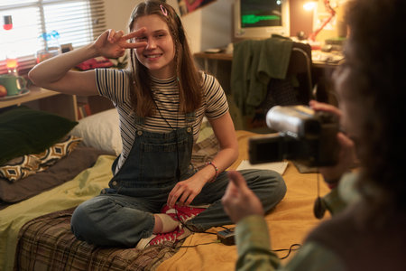 Smiling young girl sitting on bed in cozy bedroom, posing peace sign with fingers. Teenage girl being photographed in a casual indoor settingの写真素材