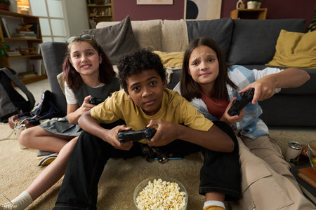Diverse group of children sitting on floor in living room, playing video games and enjoying bowl of...