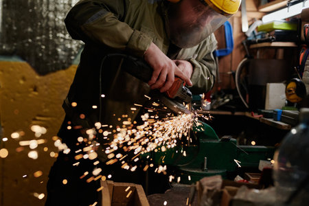 Professional metalworker in protective workwear bending over workbench with metal detail while...