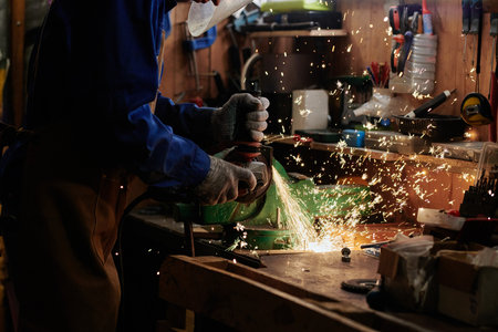 Hands of unrecognizable metalworker in protective mask and gloves grinding metal detail of...