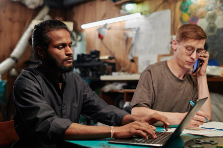 Young technician typing on laptop keyboard against colleague taking notes and talking to client on...