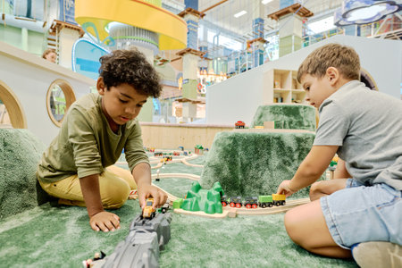 Two intercultural boys playing with toy cars and trains while sitting on the floor of playground in modern leisure centerの写真素材