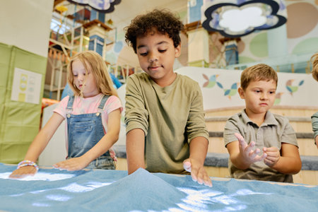 Cute African American boy and other kids drawing pictures on tabletop with blue sand in modern leisure centerの写真素材