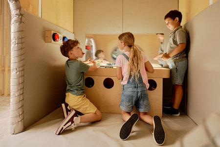 Group of three cute little children standing by table in playing room while drawing and chatting to one another or discussing ideasの写真素材